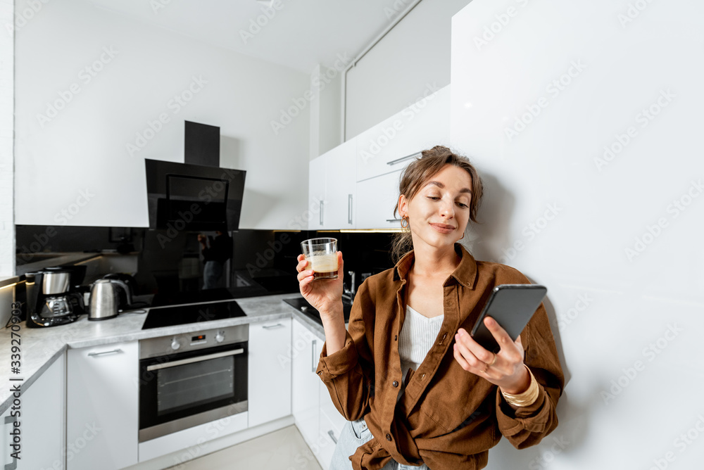 Young woman with phone and coffee on the modern kitchen at home, wide angle view. Concept of a smart