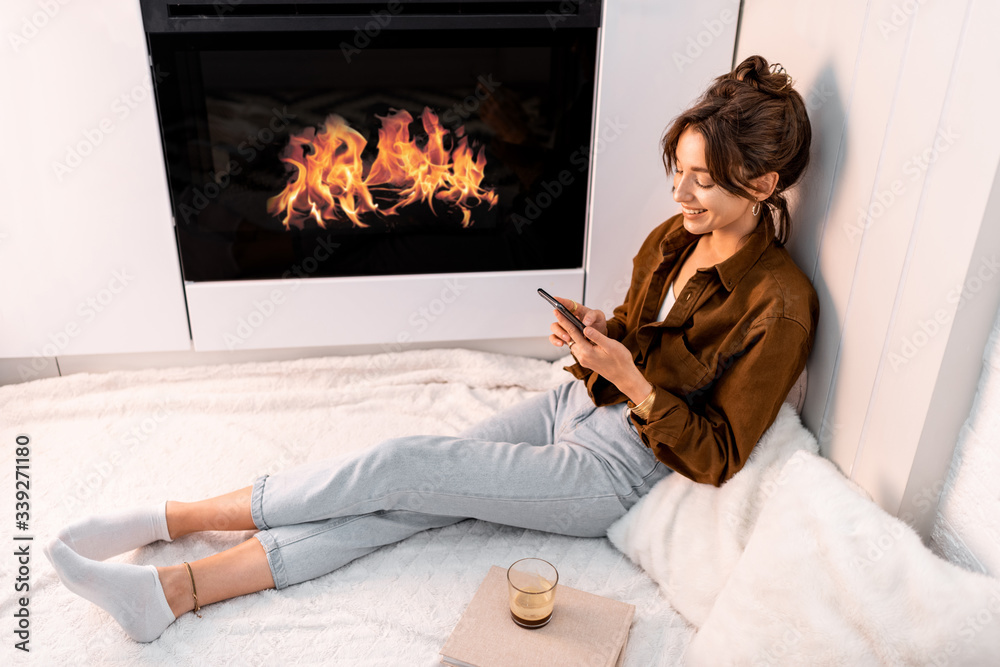 Young woman relaxing with a smart phone, sitting near the fireplace at the modern living room at hom