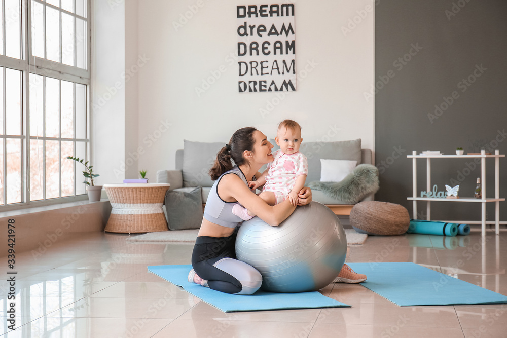 Young sporty mother and her baby doing exercises with fitball at home