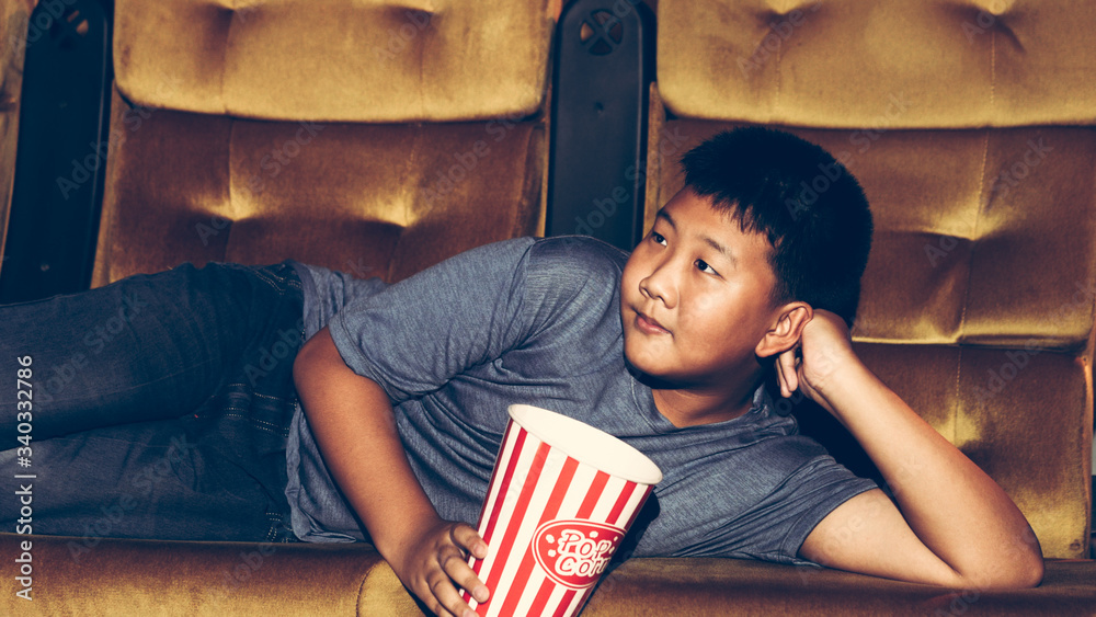 The boy is laying down watching movies and eating popcorn happily and enjoying in the cinema.