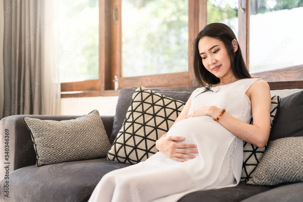 Young Asian pregnancy woman holding tummy with both hands sitting on sofa at home. Pregnant girl loo