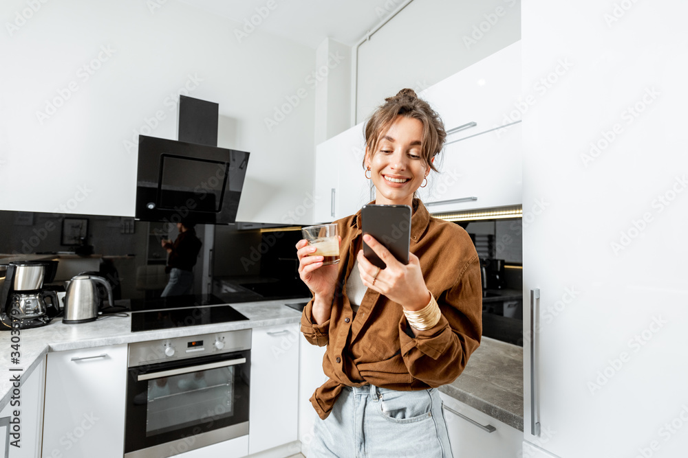 Young woman with phone and coffee on the modern kitchen at home, wide angle view. Concept of a smart