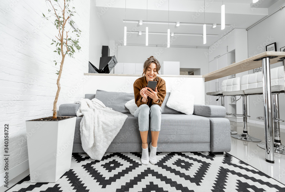 Young relaxed woman sitting with mobile phone on the couch at the living room at home, wide view on 