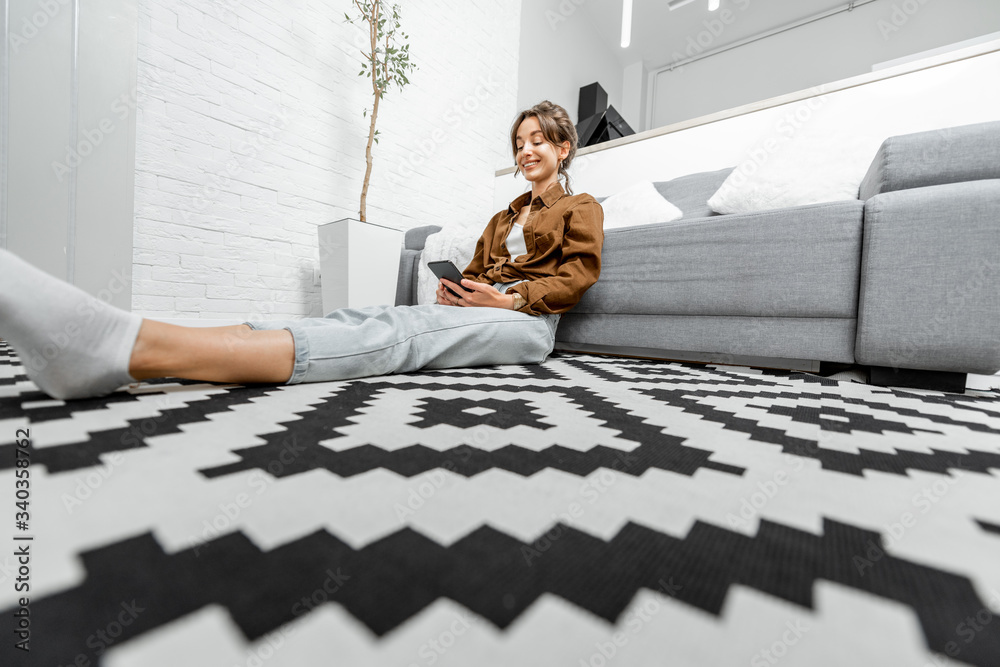 Young and cheerful woman sitting on the floor, relaxing with a smart phone at home. Wide view on the