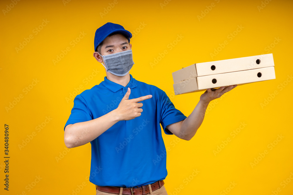 Young Asian handsome delivery man wearing mask pointing to pizza boxs over yellow isolate background