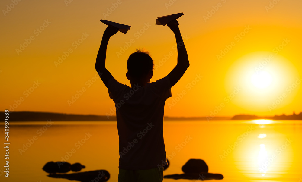 Teen boy holds paper airplanes in hands at orange sunset. A silhouette of a child with origami plane