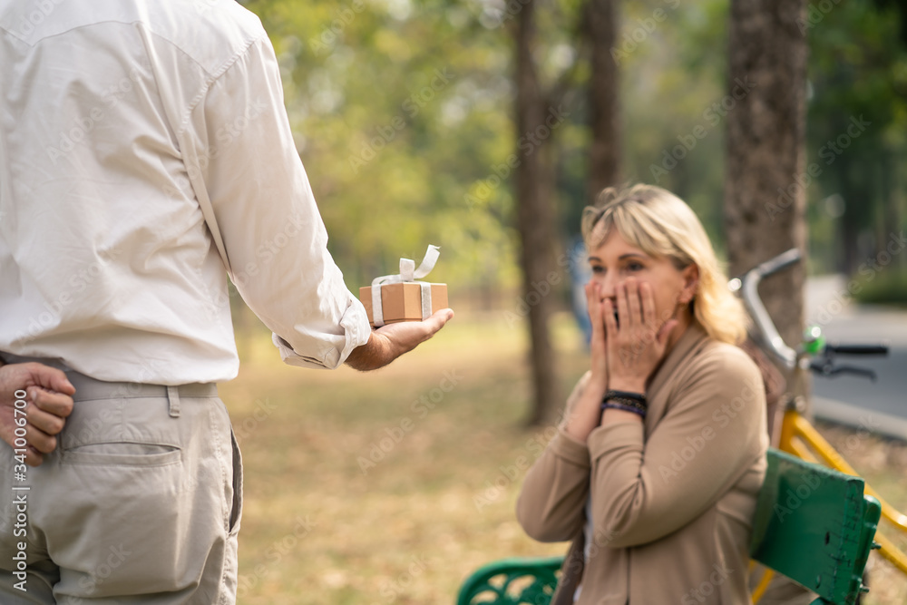 Elder senior retirement couple lifestyle in park, old man giving small gift box behind to make a sur