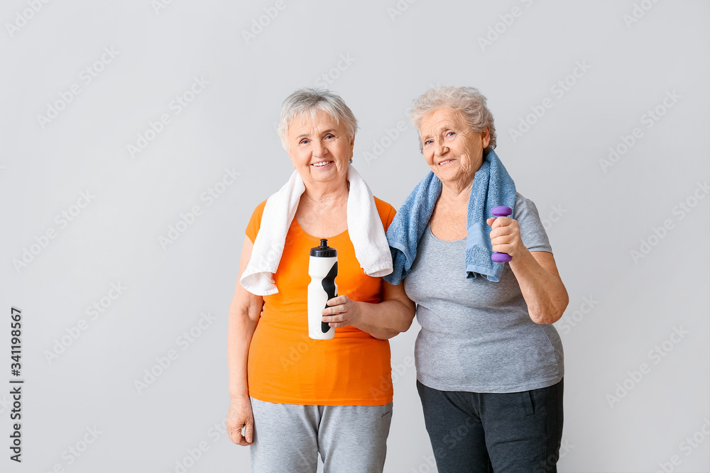 Happy elderly women with bottle of water and dumbbell on light background