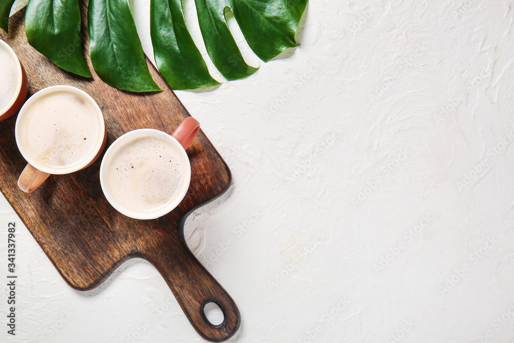 Board with cups of coffee and tropical leaf on light background