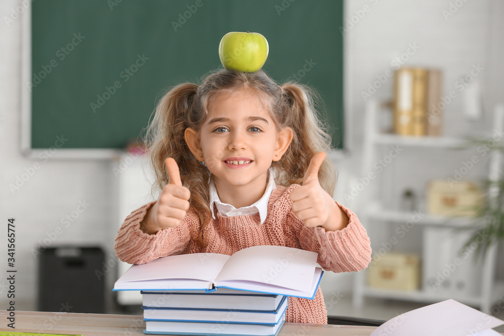 Cute little schoolgirl with books and apple showing thumb-up at desk in classroom