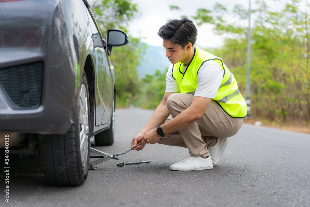 Young Asian man with green safety vest changing the punctured tyre on his car loosening the nuts wit