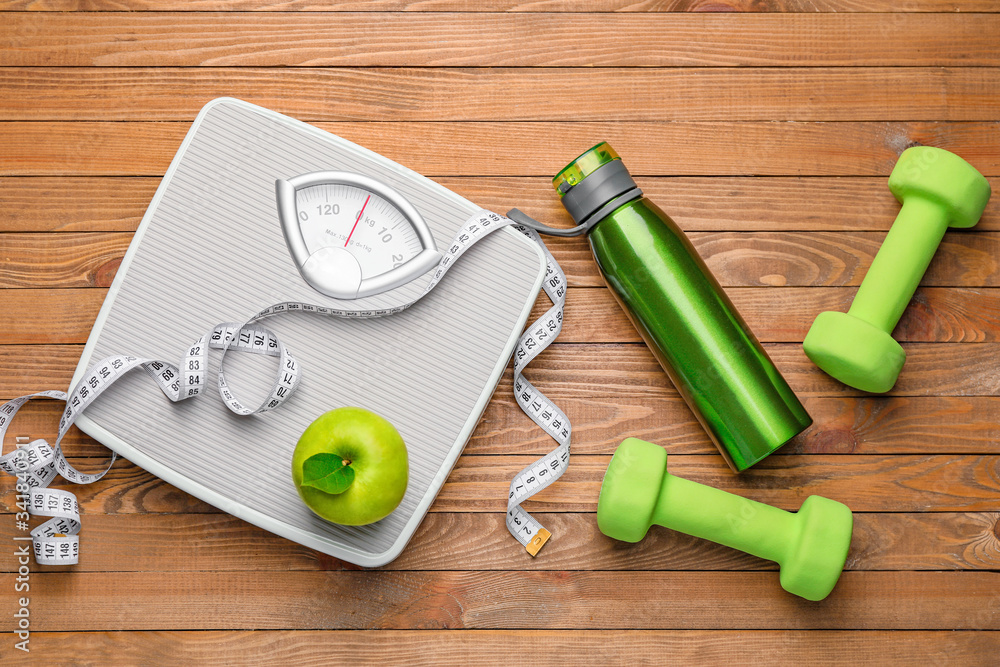 Weight scales with measuring tape, apple, bottle of water and dumbbells on wooden background