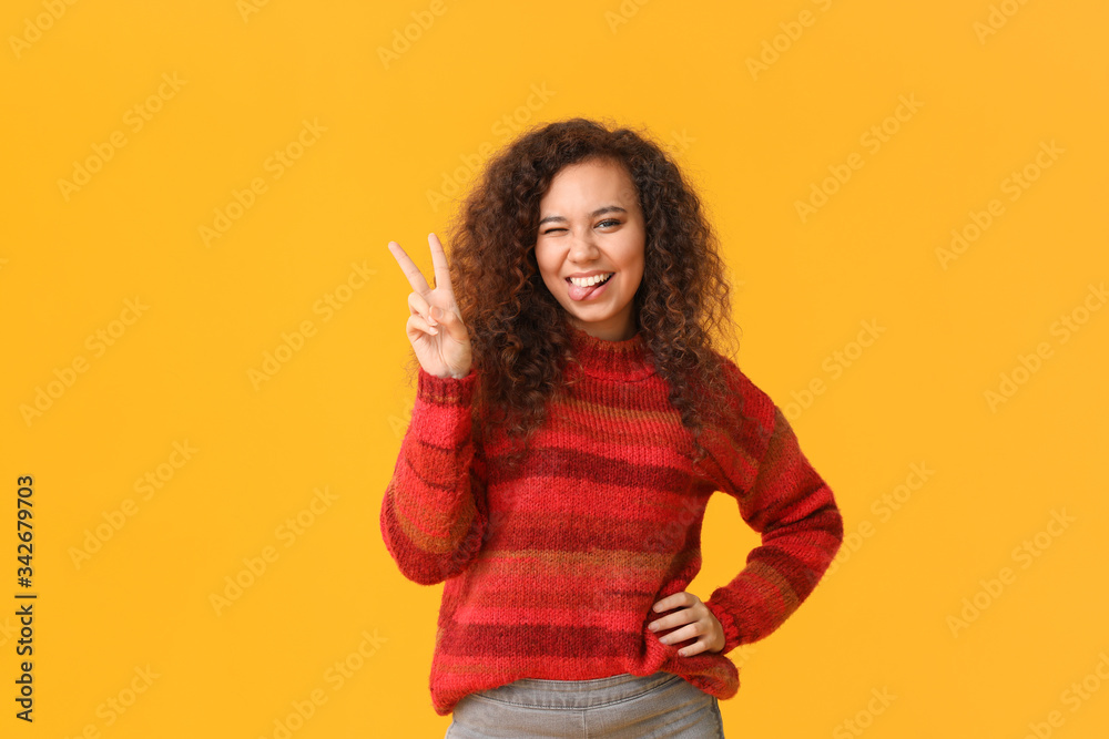 Young woman in warm sweater showing victory gesture on color background