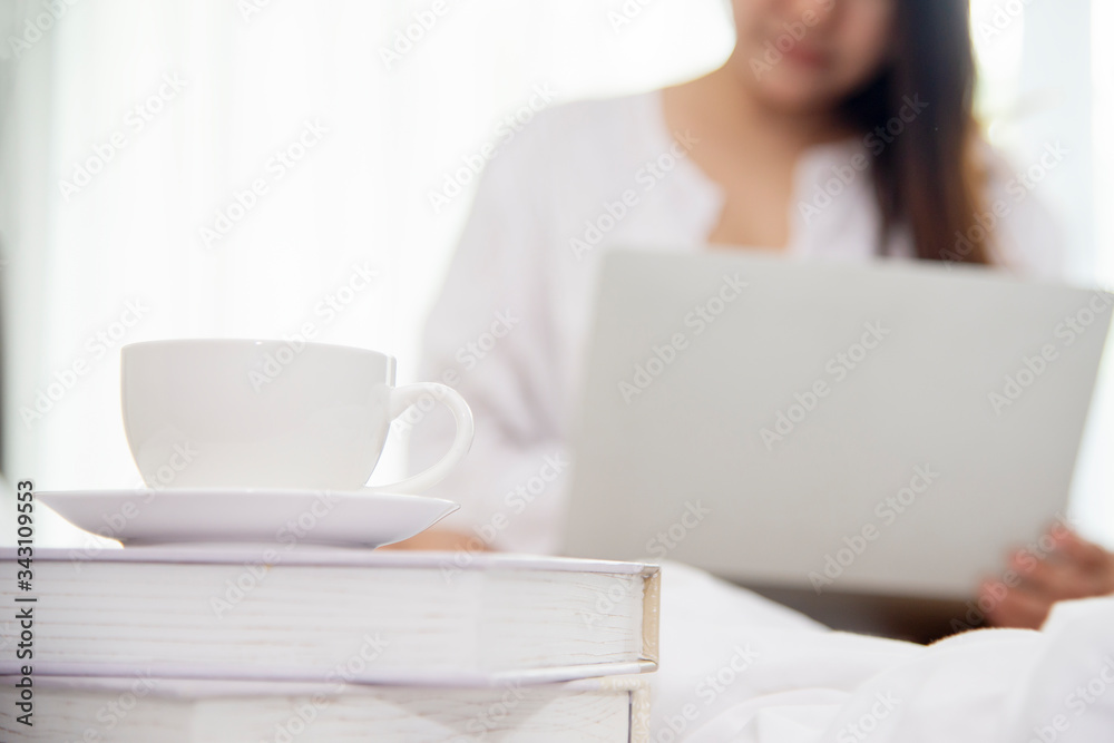 Close up on coffee cup in morning with blurred  woman sitting on bed working on laptop computer  in 