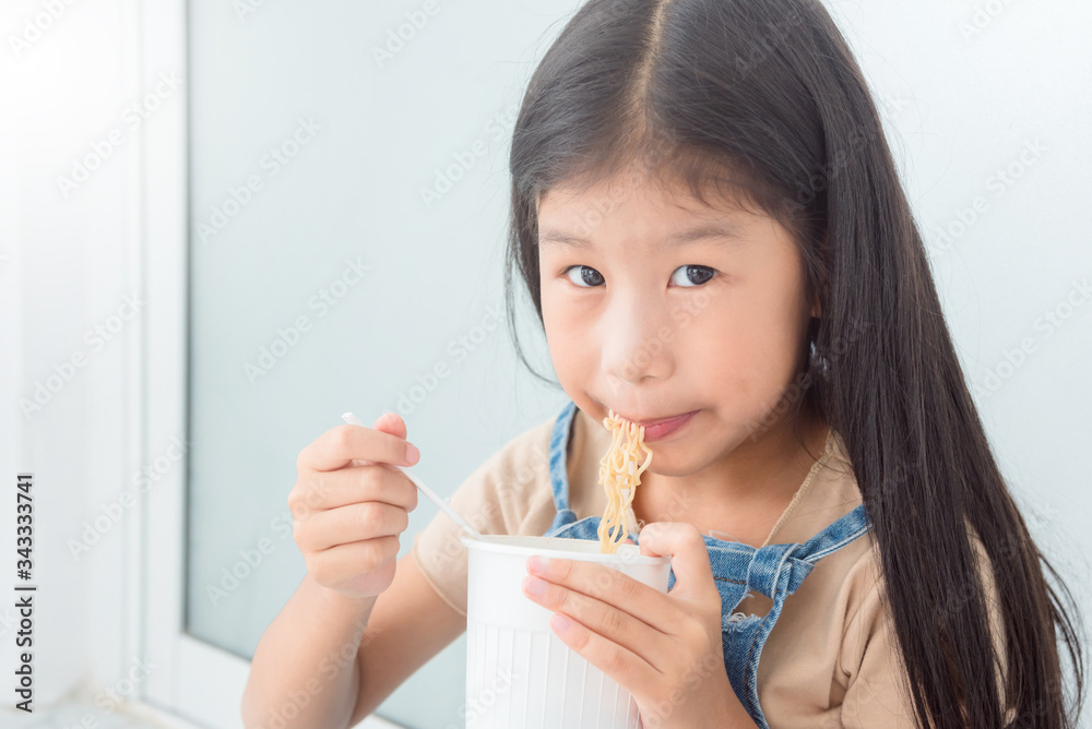 Little asian girl eating instant cup noodle by fork at home.