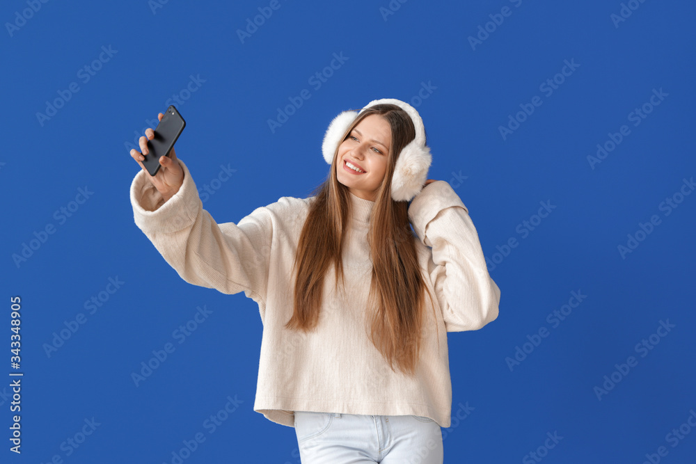 Beautiful young woman in warm sweater taking selfie on color background