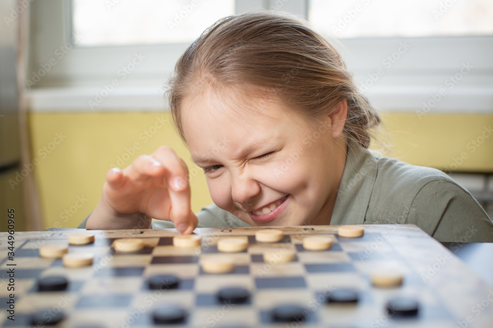 the girl playing checkers at the table closed one eye and aims at the opponent with a checker.