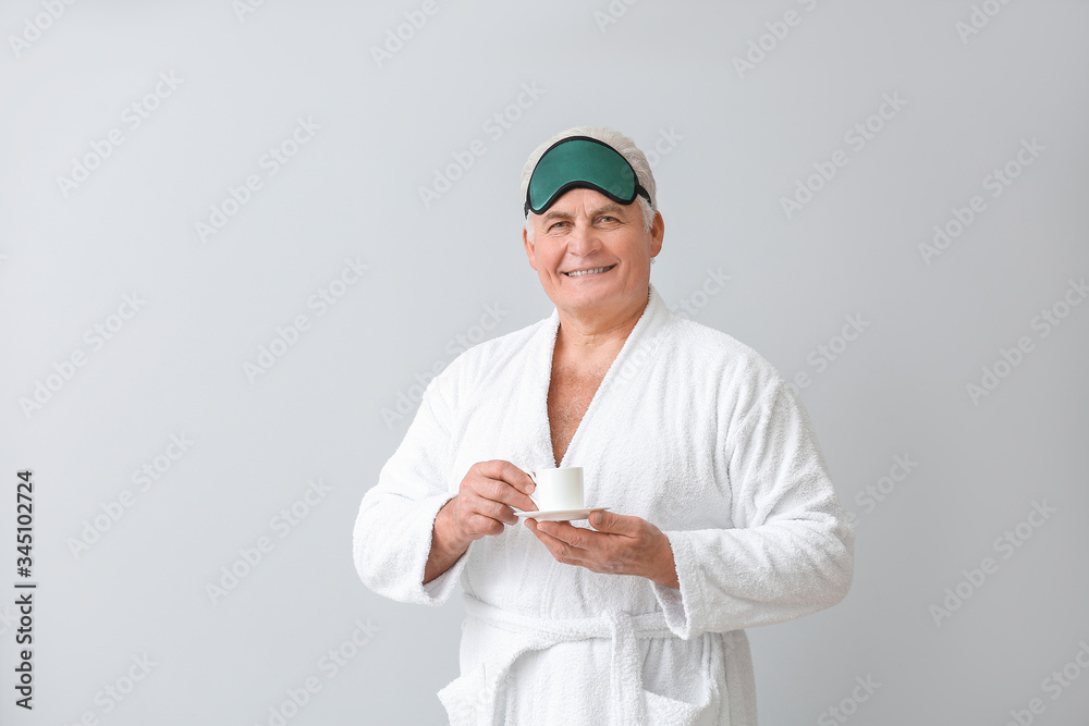 Happy mature man in bathrobe, with sleep mask and cup of coffee on grey background