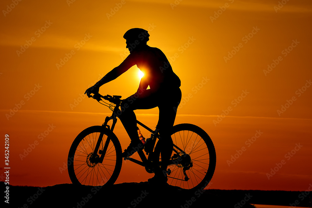 Silhouette of a biker on sky background on sunset. High hill top and wide river on orange sunset.