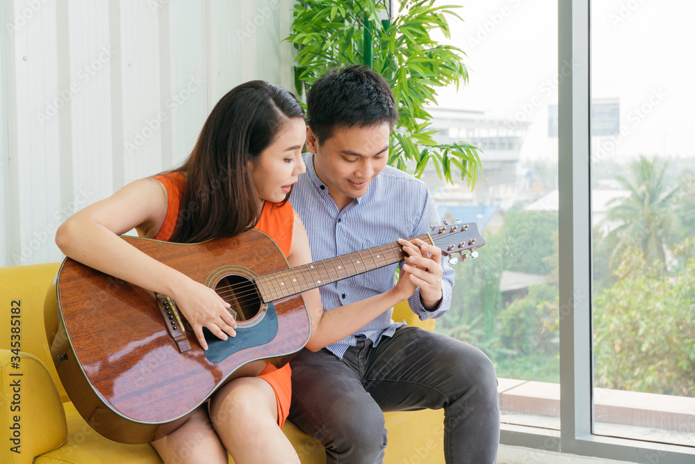 Happy smiling Asian couple in love sitting on couch while young man teaching his girlfriend playing 