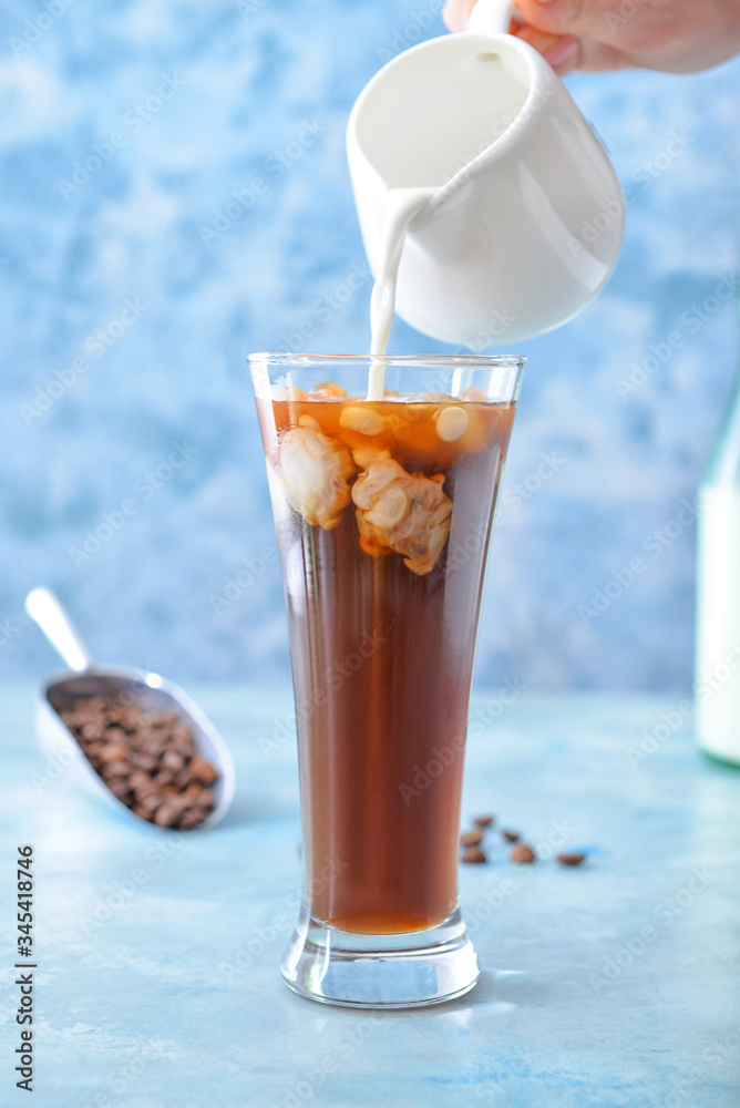 Pouring of milk into cold coffee in glass on color background