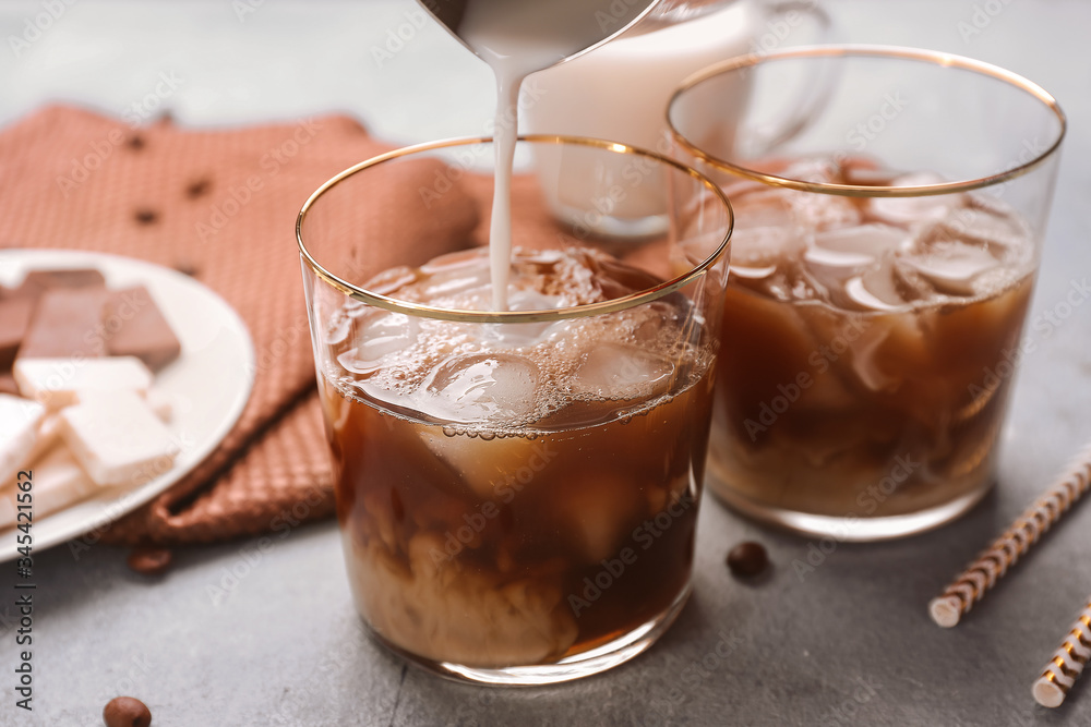 Pouring of milk into cold coffee in glass on grey background