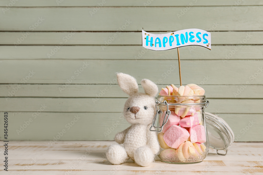 Toy bunny, jar of sweets and stick with word HAPPINESS on table