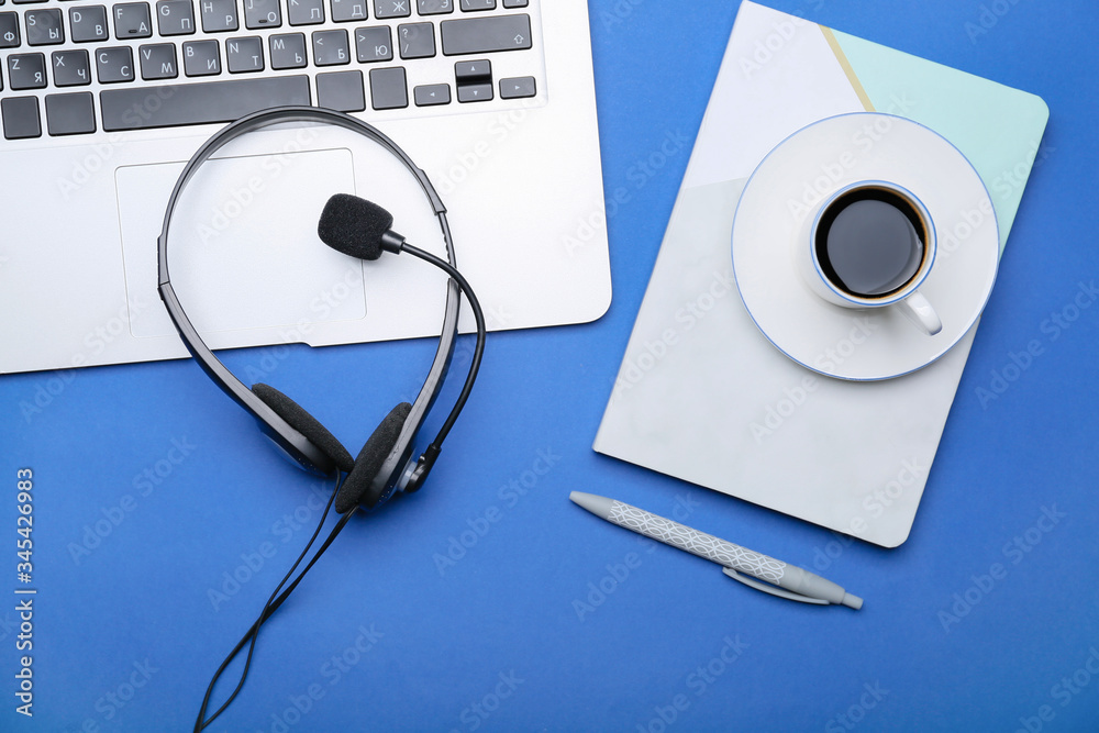 Laptop with headset, notebook and coffee on color background