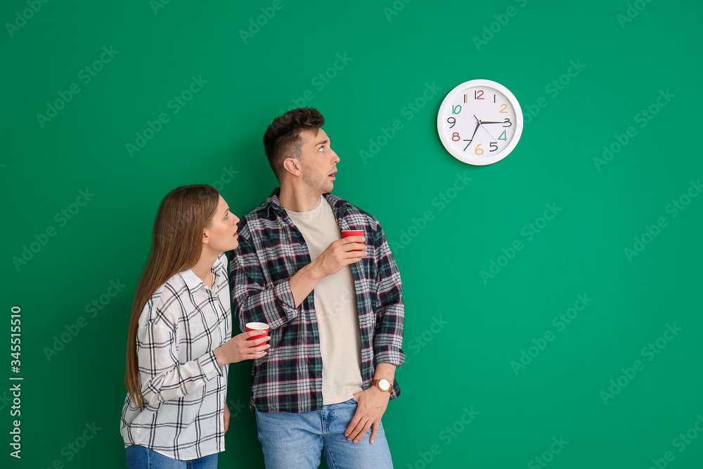 Shocked young couple with coffee looking at clock on color background