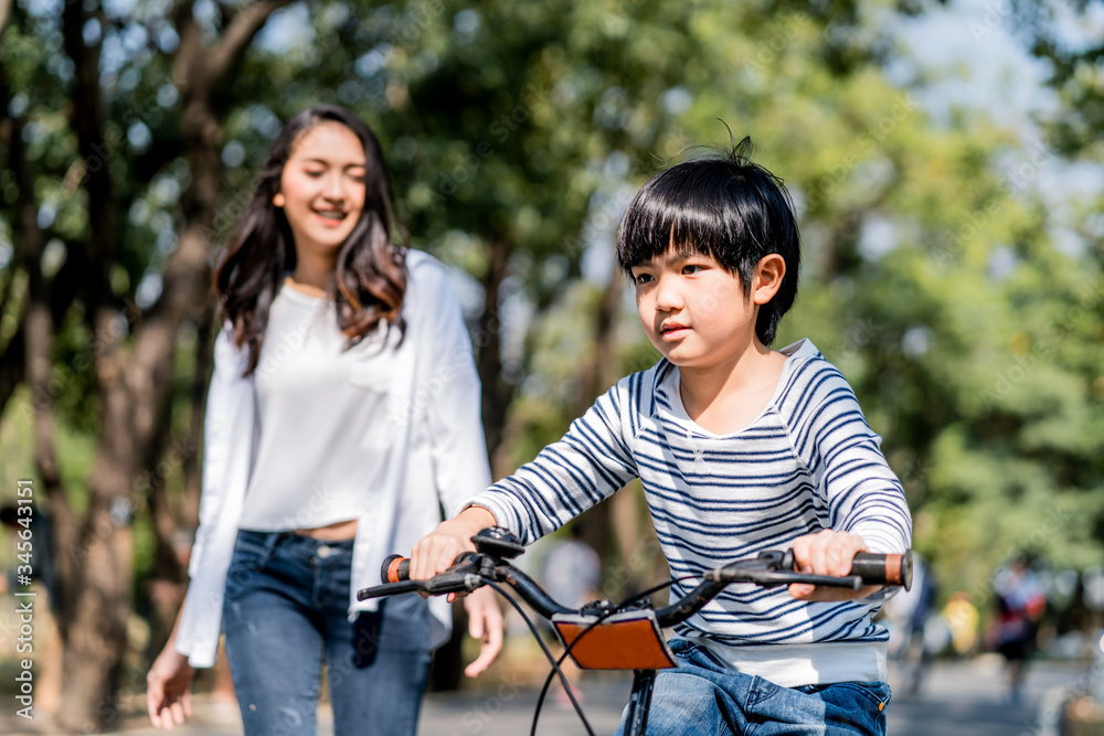 Asian young mother taking care kid while practicing to ride a bicycle by running follow son in publi