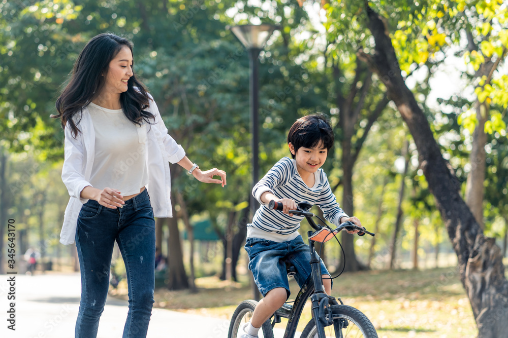 Asian young mother taking care kid while practicing to ride a bicycle by running follow son in publi