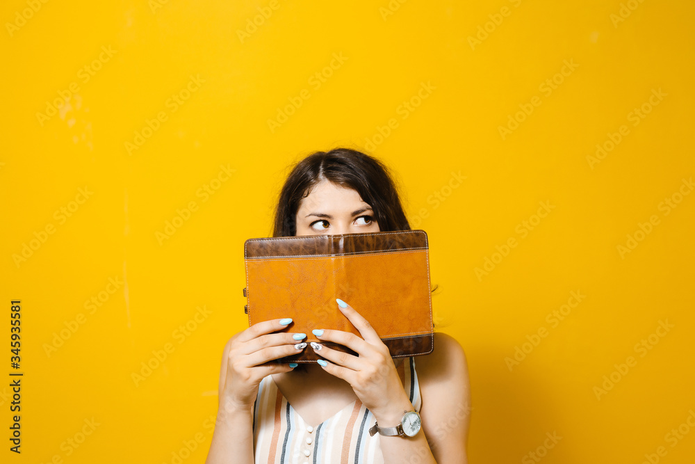 Amazed pretty young woman covered her face with book over yellow background