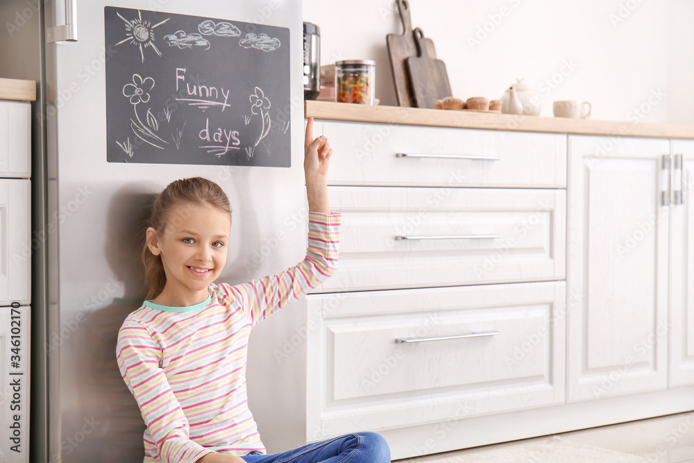 Little girl near chalkboard on refrigerator in kitchen