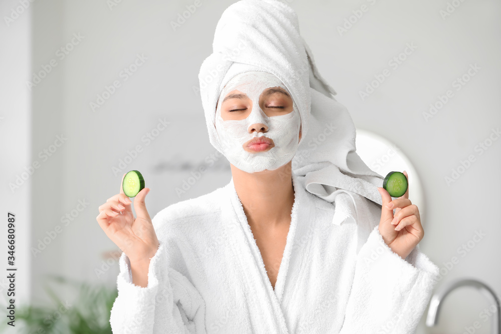 Beautiful young African-American woman with cucumber slices and facial mask in bathroom