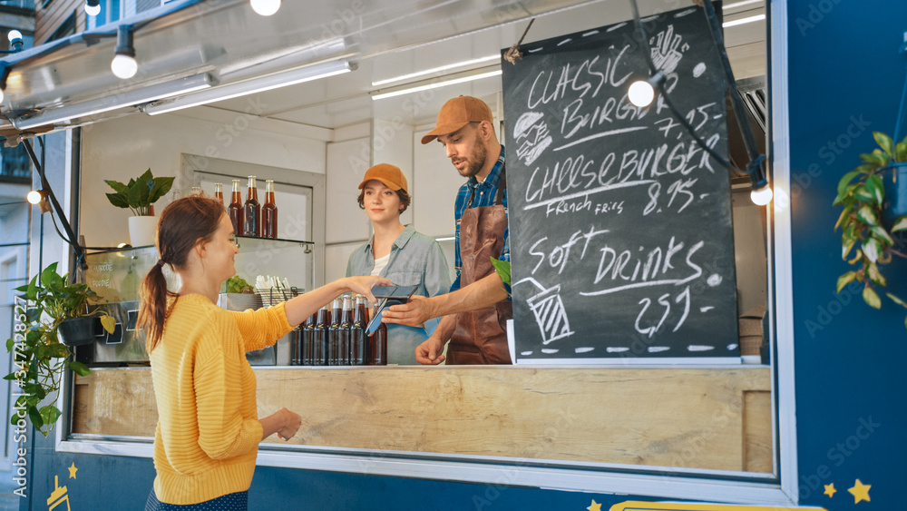 Food Truck Employee Hands Out a Freshly Made Burger to a Happy Young Female. Young Lady is Using NFC