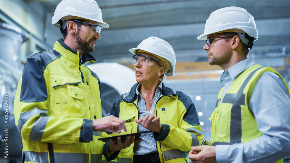 Three Heavy Industry Engineers Stand in Pipe Manufacturing Factory, Use Digital Tablet Computer, Hav