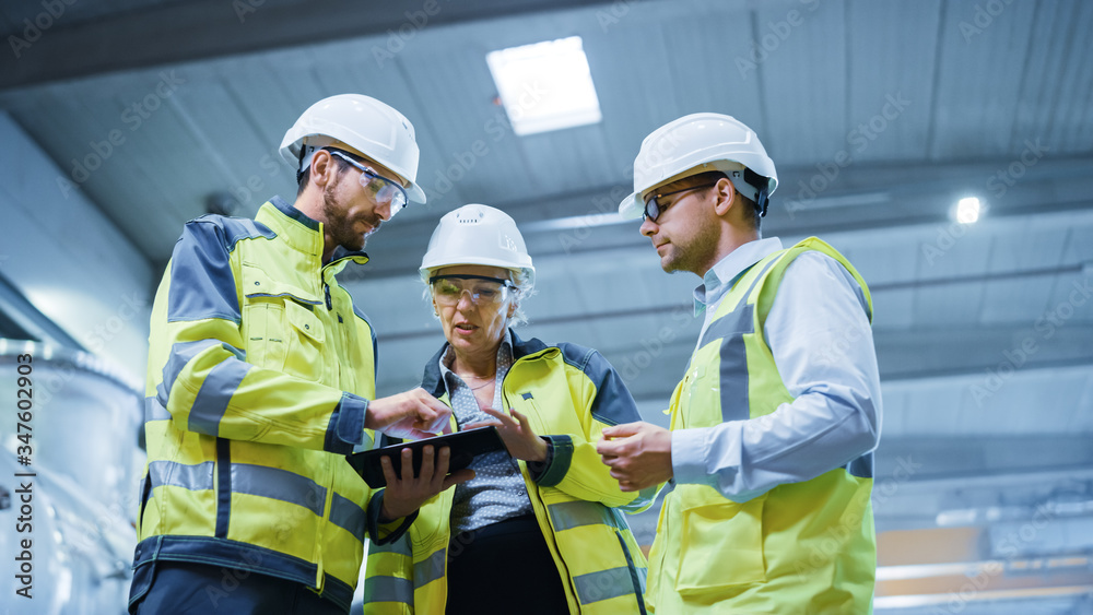 Three Heavy Industry Engineers Stand in Pipe Manufacturing Factory, Use Digital Tablet Computer, Hav