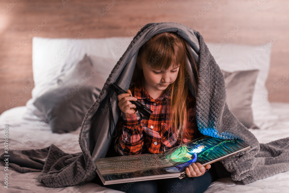 Little girl with flashlight reading book in evening