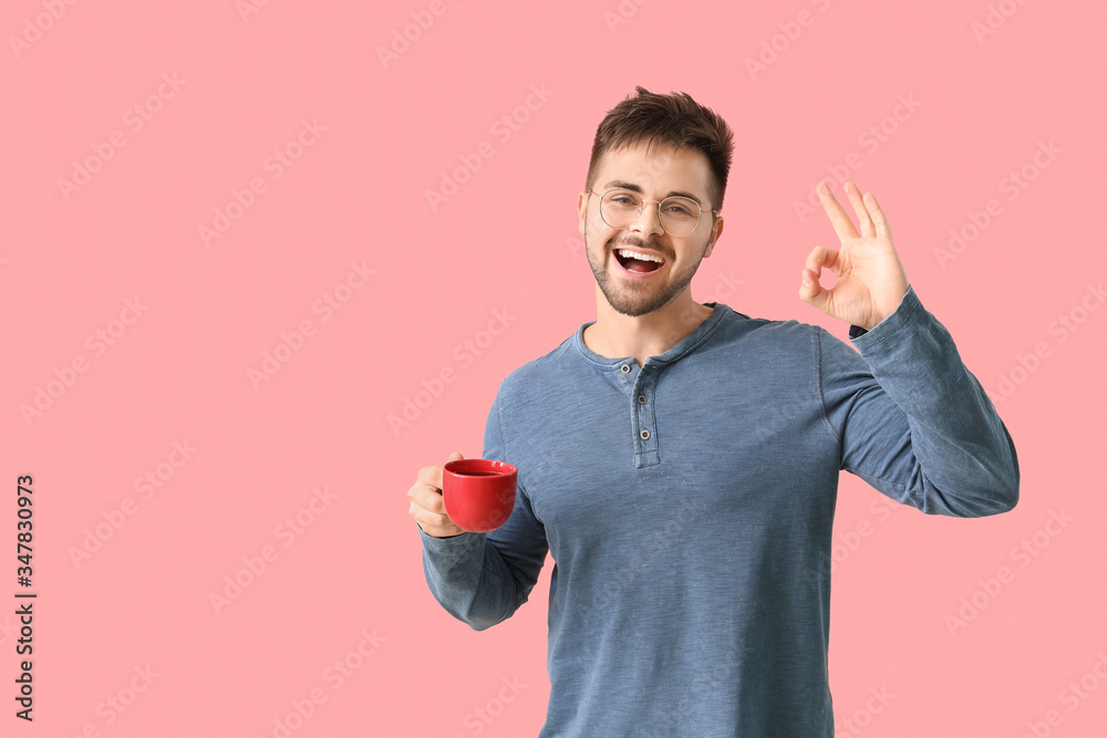 Young man with cup of hot coffee showing OK on color background