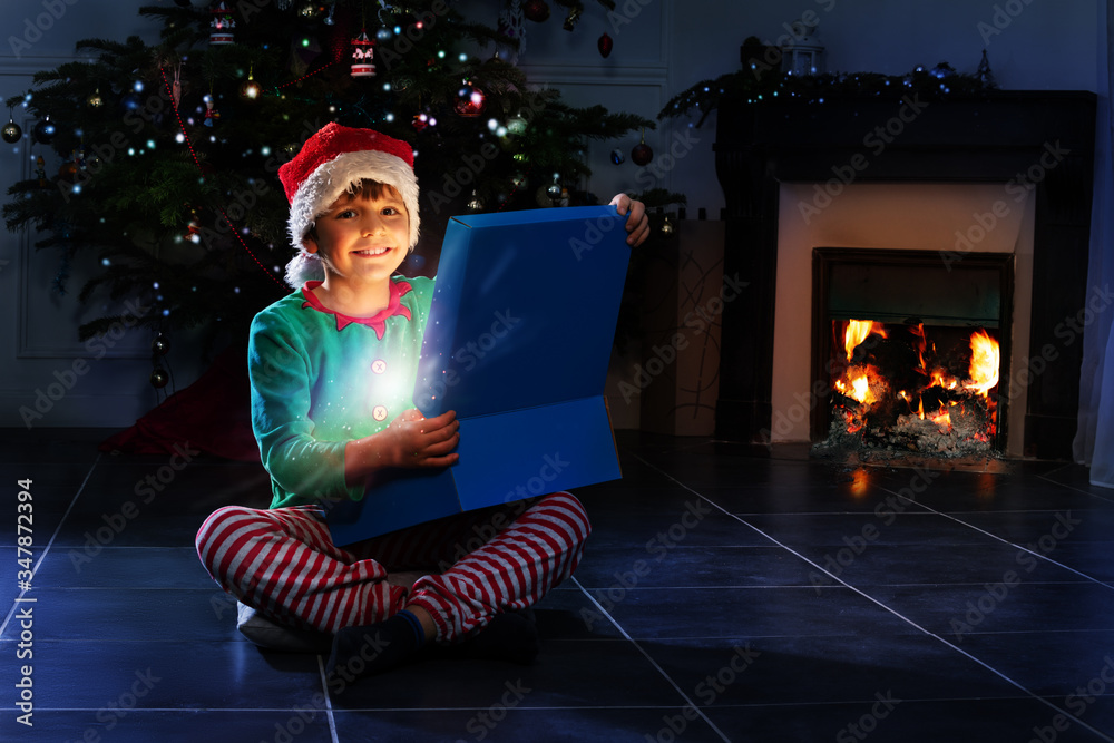 Happy little boy opens shining Christmas present box sitting near fireplace