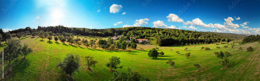 Wide aerial panorama view of a nice rural landscape with green meadows on hills, trees and a few hou