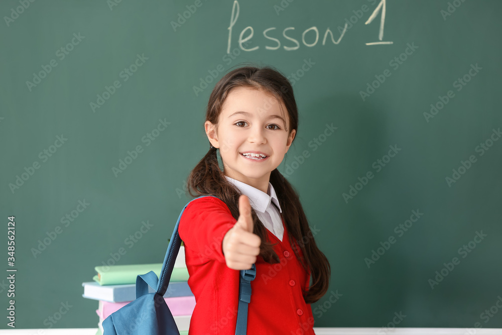 Cute little pupil showing thumb-up near blackboard in classroom