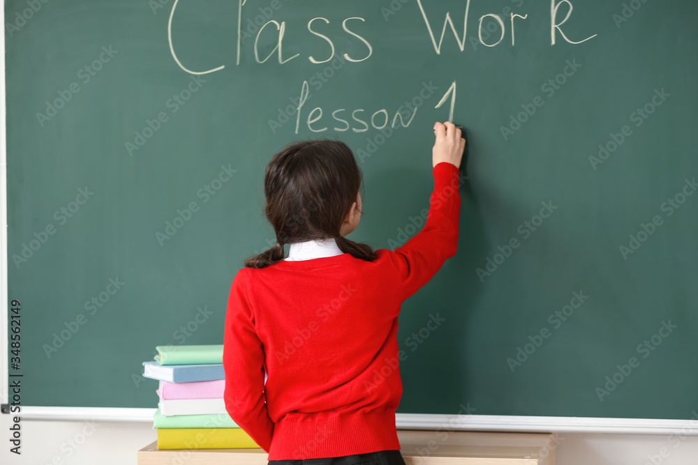 Cute little pupil writing on blackboard in classroom