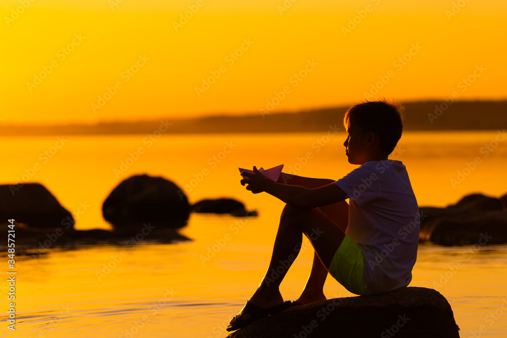 Little boy with paper ship in hands. Beautiful orange sunset. Origami. River. Lake.