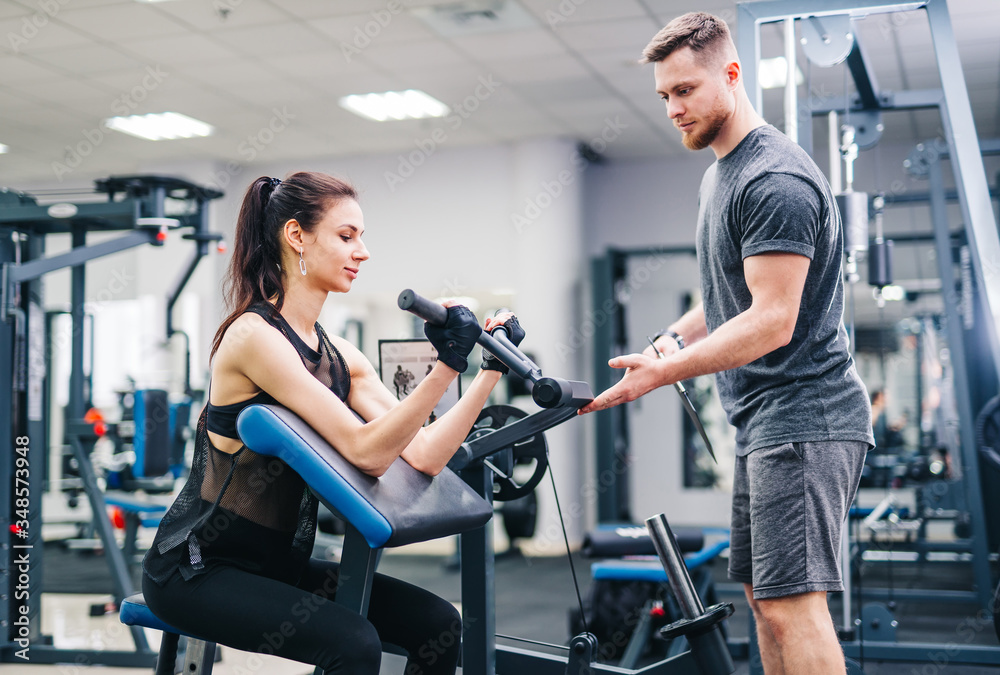 Brunette fitness girl trains in a gym with a barbell disk. Trainer helps achive better results.