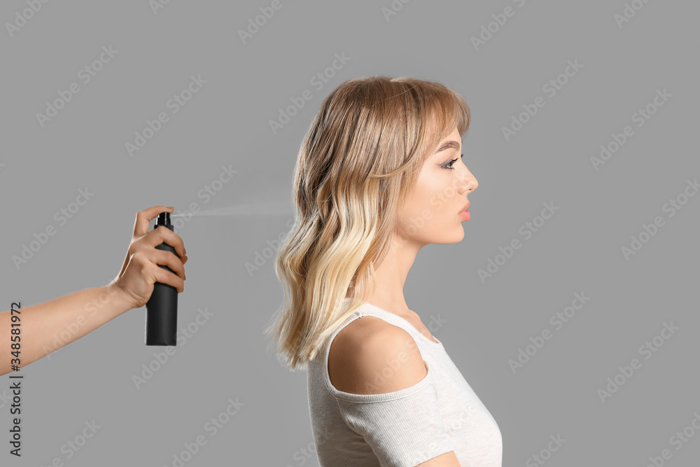 Beautiful young woman and hand with hair spray on grey background