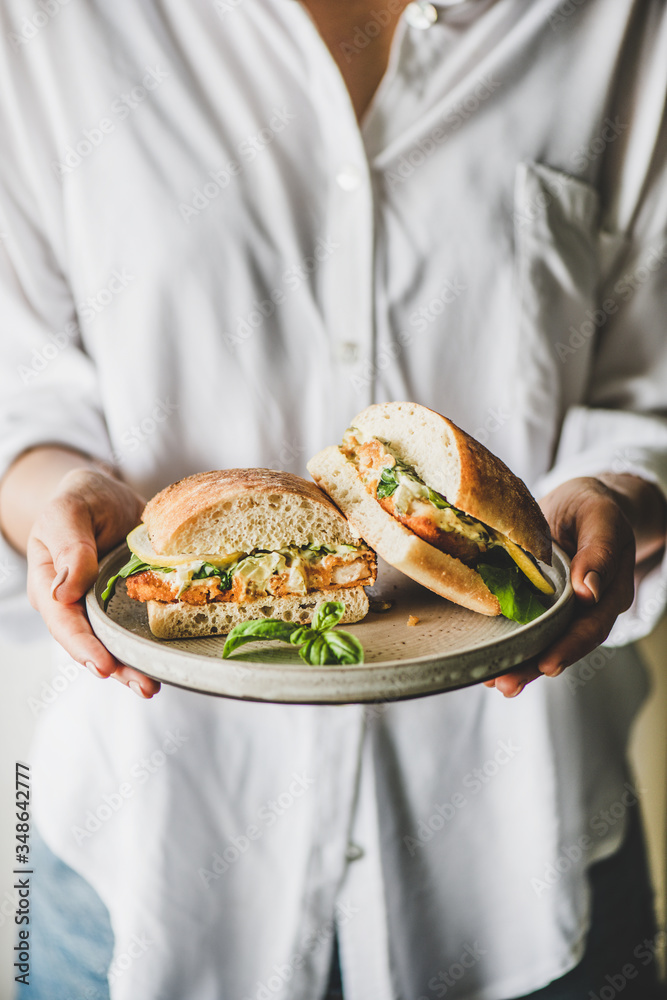 Woman in white shirt holding plate with fresh fried fish sandwich with tartare sauce, lemon and arug