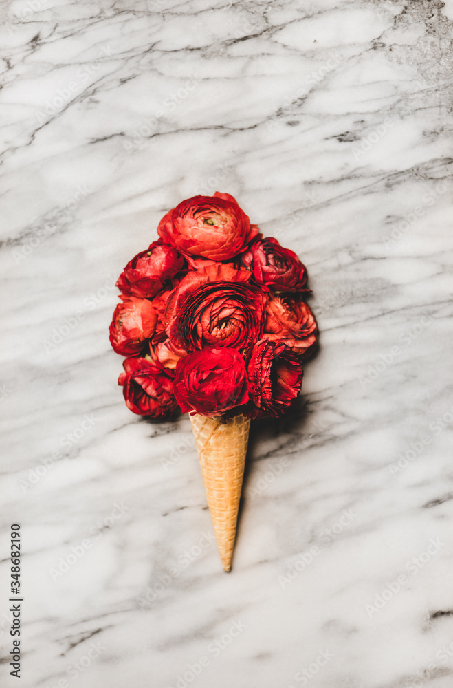 Flat-lay of ice cream waffle cone with scoop of red blooming ranunculus flowers over white marble ta