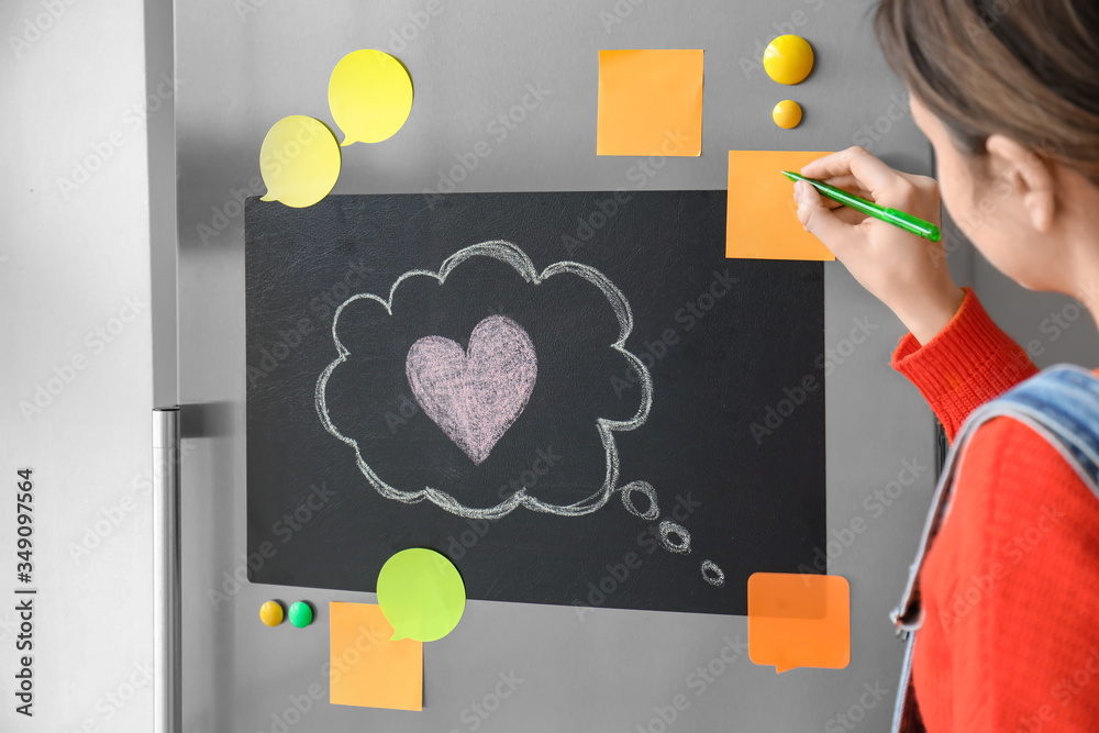 Young woman writing on sticky paper attached to refrigerator door