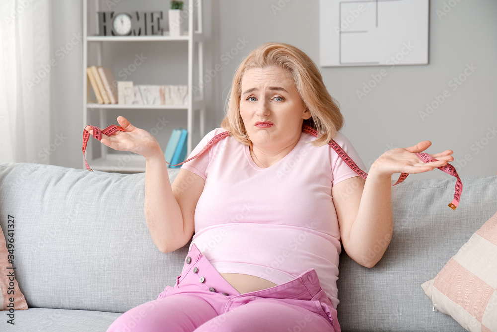 Sad overweight woman in tight clothes and with measuring tape at home